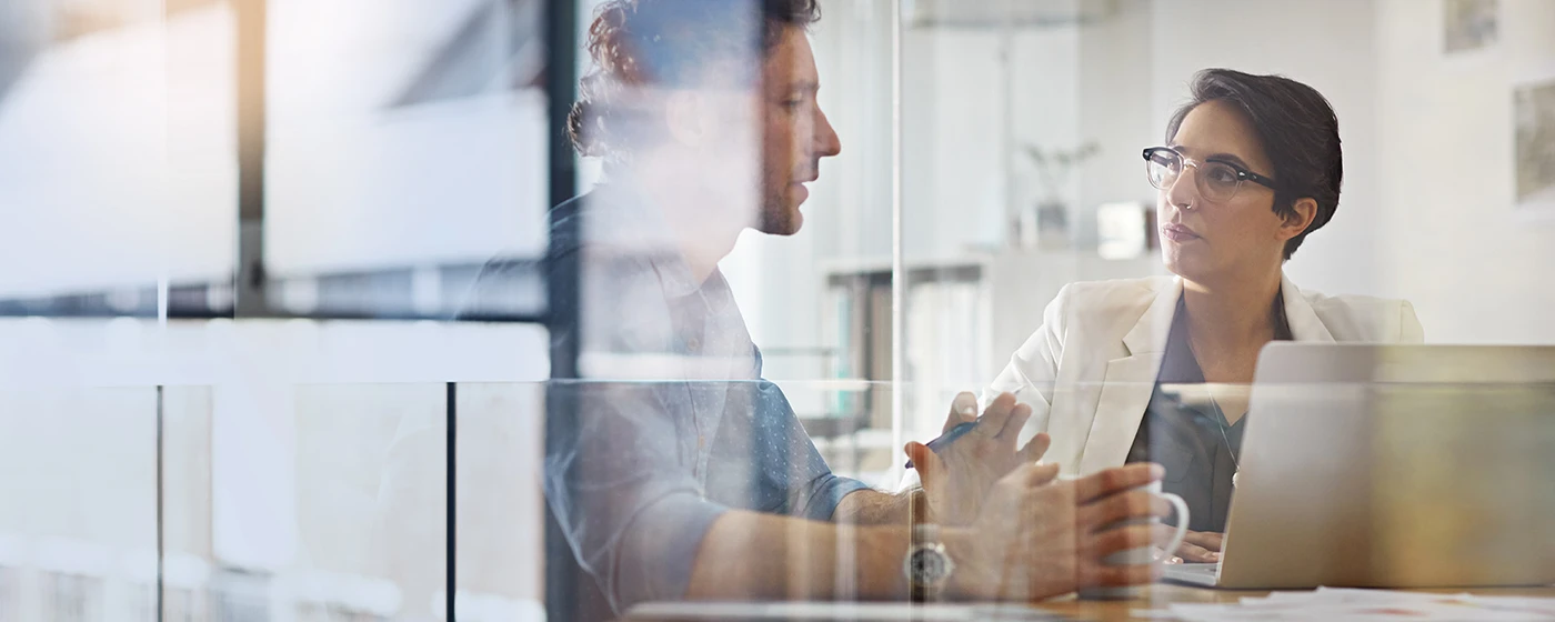 Mitarbeiter erklärt einer Mitarbeiterin etwas hinter einer Glasscheibe
